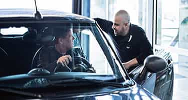 MINI Service Technician in service bay leaning on MINI vehicle speaking with male driver.